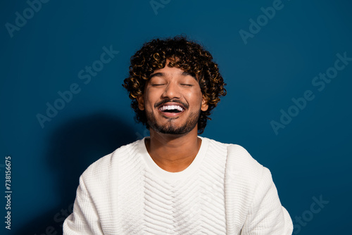 Portrait of young funny guy in white jumper curly hair student from mexico laughing closed eyes isolated on dark blue color background © deagreez