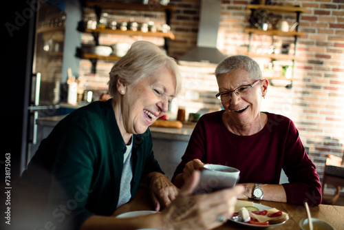 Excited senior woman sharing smartphone with a friend over coffee at home