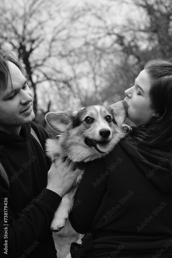 A young man and woman are hugging with their cute Welsh corgi puppy in the park. Dog with happy expression on its face. Life style portrait. Trendy monochrome. A young man and woman are hugging with their cute Welsh corgi puppy in the park. Dog with happy expression on its face. Life style portrait. Trendy monochrome.