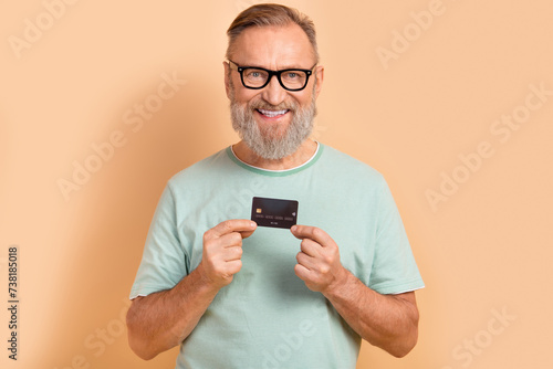 Photo of satisfied pensioner person in eyewear dressed teal t-shirt showing credit card in arms isolated on beige color background © deagreez