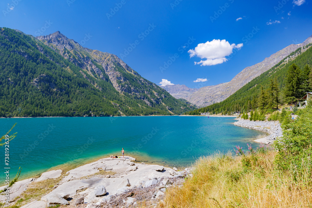 Obraz premium Ceresole Reale, Italy. View of Lake Ceresole on a sunny summer day. Some people sunbathing on the lake shore. Alps mountains in the background. Mountain landscape. 2023-08-20.