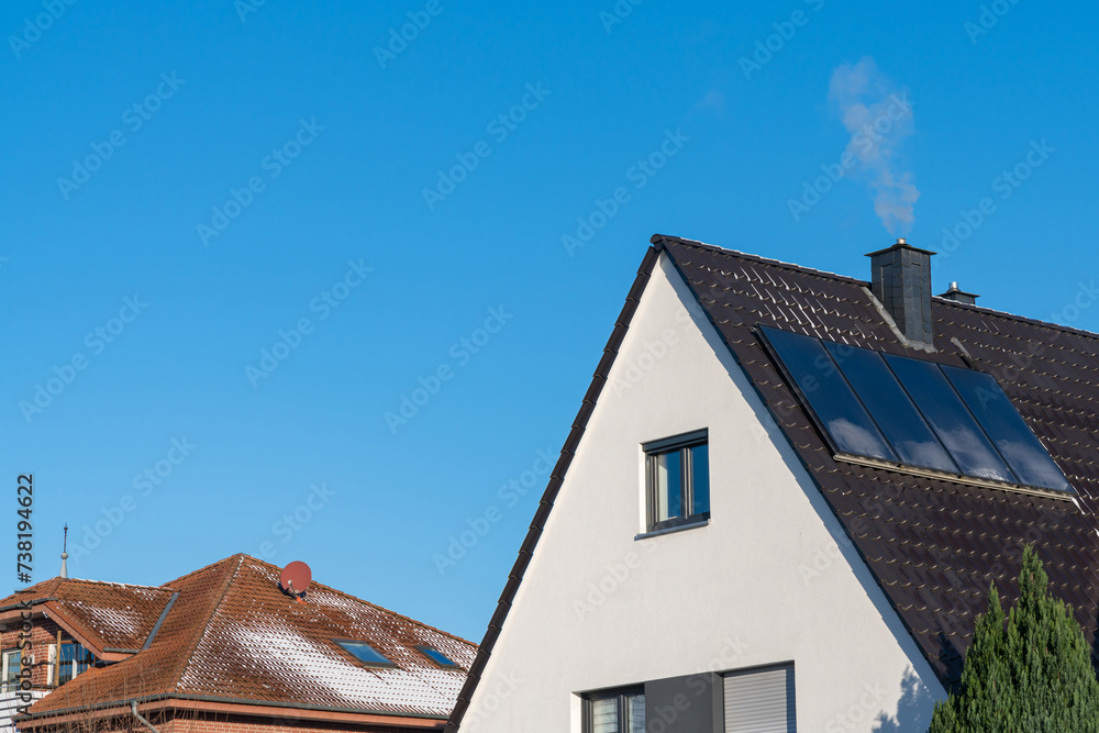 Light smoke from a chimney on a tiled roof with solar panels. Clear blue sky.