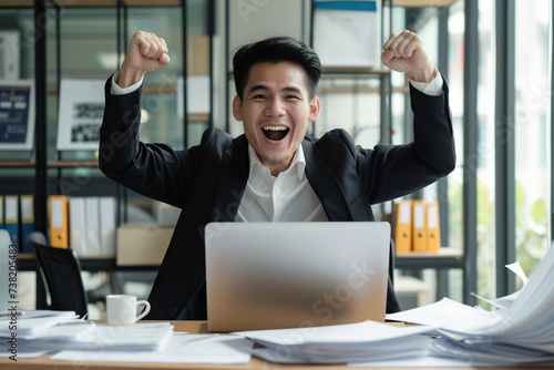 Happy excited young Asian business man accountant standing at the desk working on laptop computer with a pile of documents on table in office and making yes gesture rejoicing in successful job.