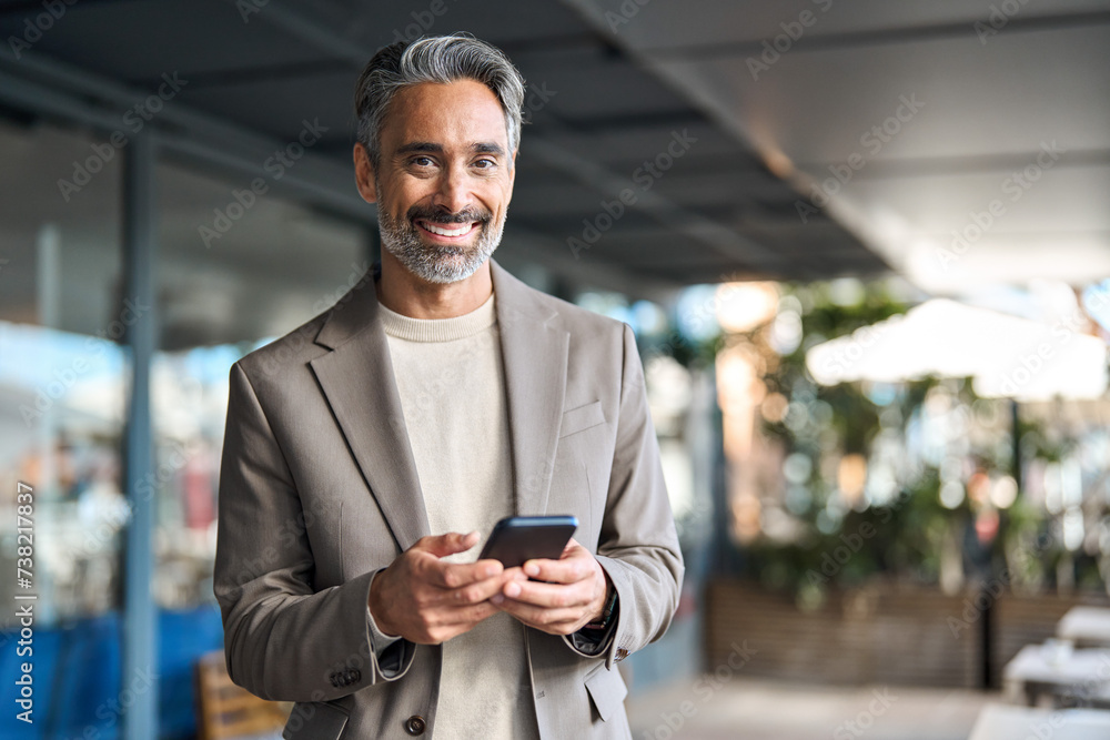 Happy mature older business man executive using mobile cell phone outdoors. Middle aged businessman manager or entrepreneur holding smartphone looking at camera standing outside office. Portrait. Happy mature older business man executive using mobile cell phone outdoors. Middle aged businessman manager or entrepreneur holding smartphone looking at camera standing outside office. Portrait.