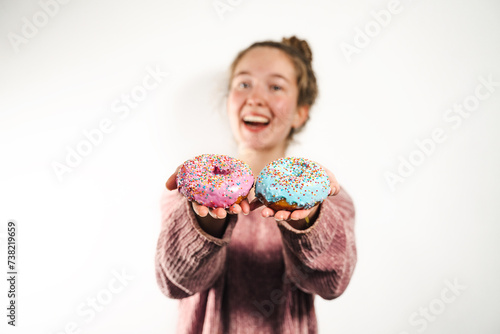 Young happy sweet tooth woman holding out a pink and blue glazed donut to the camera, tasty food and diet, high-calorie, sweets, junk food, fast sood, cheat meal day, breaking diet, foreground focus photo