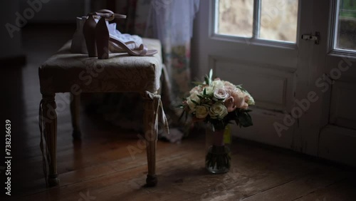 Imagen estática de zapatos de novia sobre una silla, un ramo de flores en un florero en el suelo frente a un ventanal