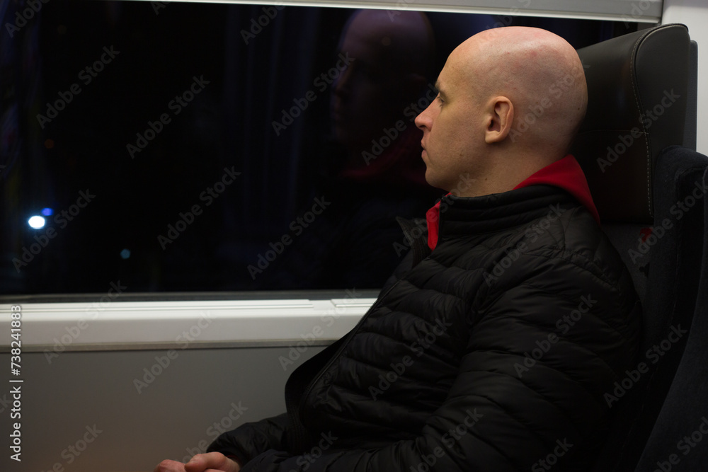 Man without hair sits near the window in a sedentary train during a trip to another city, traveling at night, a bald man near the train window Man without hair sits near the window in a sedentary train during a trip to another city, traveling at night, a bald man near the train window
