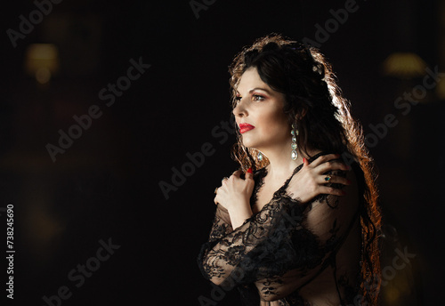 A young sexy woman in black lingerie stands alone in a dark room.