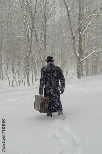Retro Man walking away in snow carrying vintage suitase wearing long coat boler hat