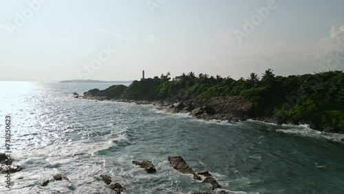 Aerial view captures lighthouse atop hill, ocean waves crash against rocky shore. Maritime beacon guides ships, scenic landmark amidst vibrant flora, overlooks vast sea, symbol of safe harbor. photo