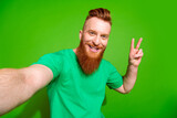 Portrait of pleasant cool guy with long red beard wear stylish t-shirt making selfie show v-sign isolated on green color background