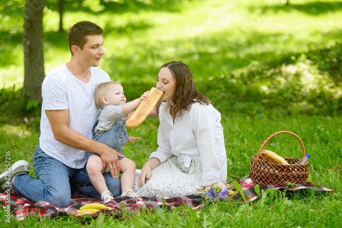 Dad, mom and daughter are sitting in a meadow on a blanket in the park on a warm sunny day,child holds a baguette in his hands, treats his mother. Family weekend getaway