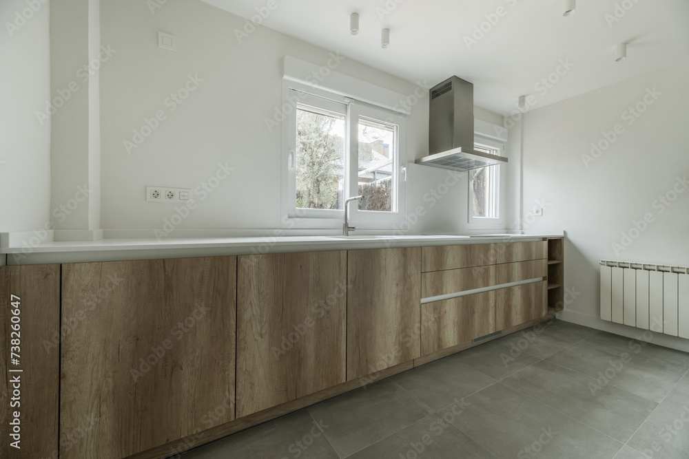 Image of a kitchen with wooden furniture, white synthetic stone countertop, stainless steel extractor hood, several windows and white aluminum radiator Image of a kitchen with wooden furniture, white synthetic stone countertop, stainless steel extractor hood, several windows and white aluminum radiator