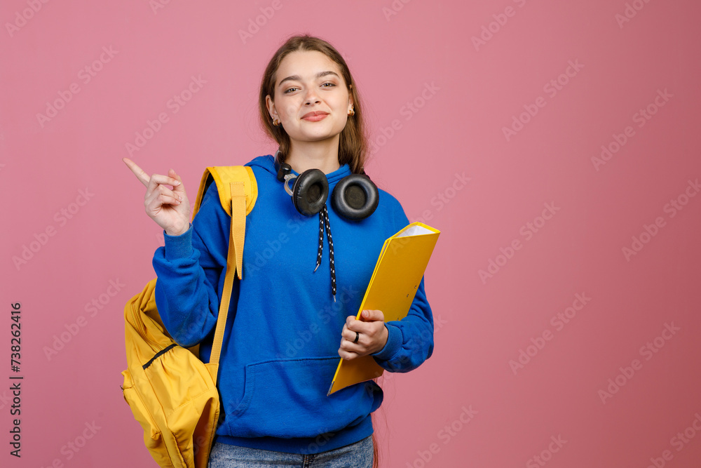 Front view of pretty brunette female standing, pointing by finger. Beautiful schoolgirl looking at camera, smiling, holding yellow rucksack and folder. Concept of youth life.