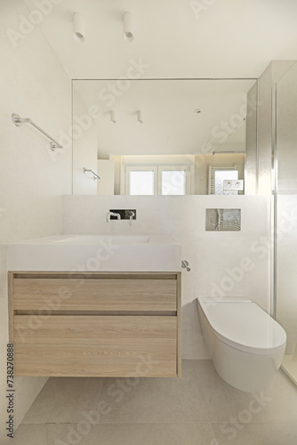 Frontal image of a small modern bathroom with an elongated white stone imitation sink with a wooden cabinet underneath and a hanging toilet