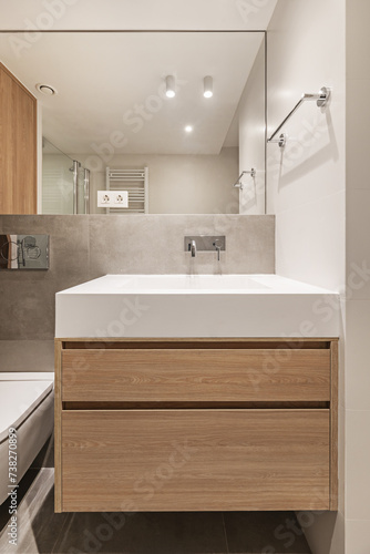 Frontal image of a small modern bathroom with an elongated white stone imitation sink with a wooden cabinet with drawers underneath  a mirror integrated into the wall and a heated towel rail