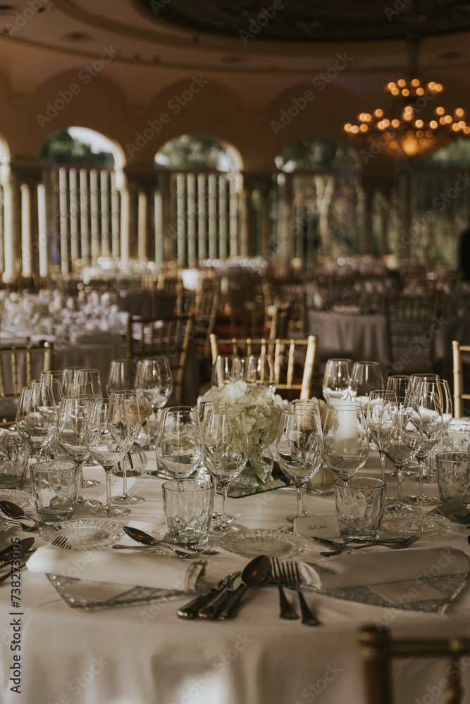 
Salón de celebraciones repleto de mesas decoradas en tonos blancos y sillas doradas. Mesas redondas con manteles blancos, cubertería y vasos de cristal transparente.