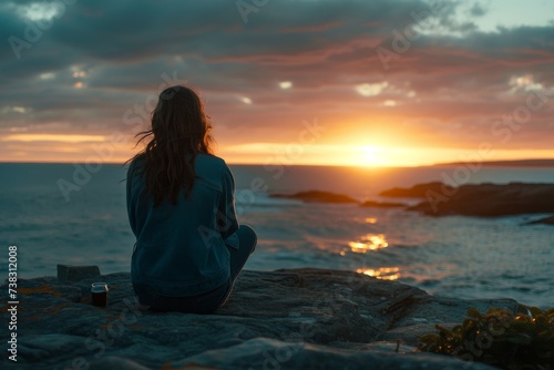 A young woman sitting on a rock by the ocean, praying as the sun sets behind her, creating a picturesque harmony of human and nature.