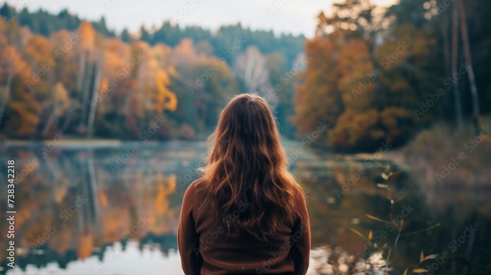 In the tranquil autumn forest, a woman gazes at her own reflection in the still waters of the lake, her hair blowing in the crisp breeze - obrazy, fototapety, plakaty 