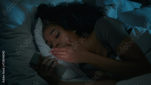 Sleepy African American woman tired girl sleep nap rest at night home dark bedroom sleeping in bed with mobile phone scrolling smartphone addict gadget read social media yawning fall asleep nighttime photo