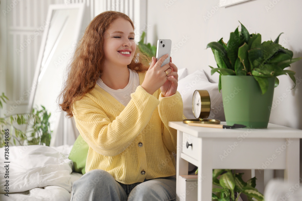Young woman with mobile phone taking picture of plant in bedroom Young woman with mobile phone taking picture of plant in bedroom