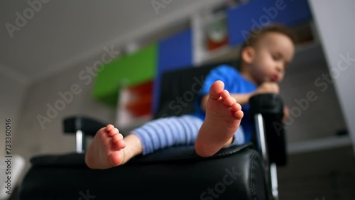 Toddle sitting in the chair his bare feet close to camera. Little child leans in the chair looking down. Blurred backdrop. Low angle view. photo