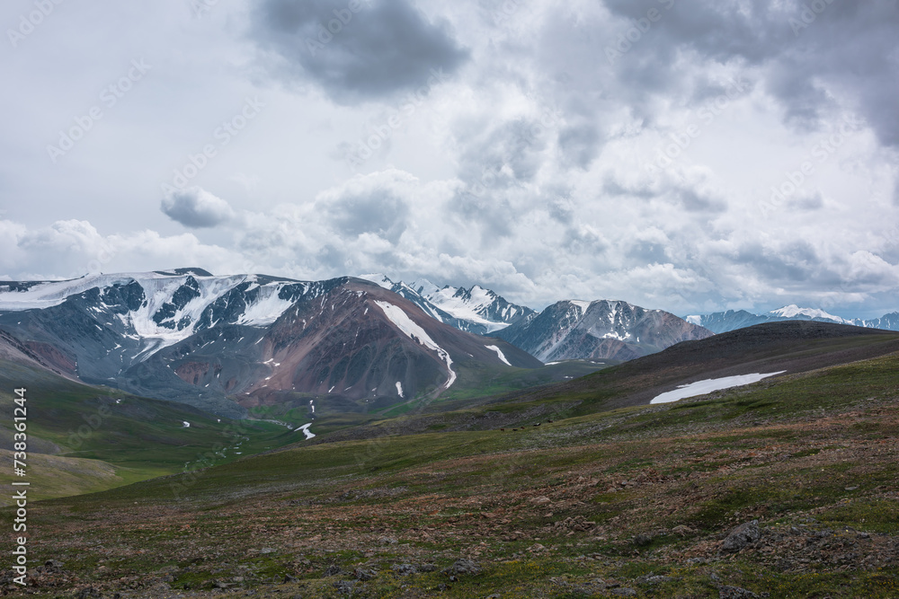 Dramatic alpine view from stony grassy hill to large snow-capped mountain range under cloudy gray sky. Beautiful mountain landscape with green rock hills against high snowy mountains in rainy weather. Dramatic alpine view from stony grassy hill to large snow-capped mountain range under cloudy gray sky. Beautiful mountain landscape with green rock hills against high snowy mountains in rainy weather.