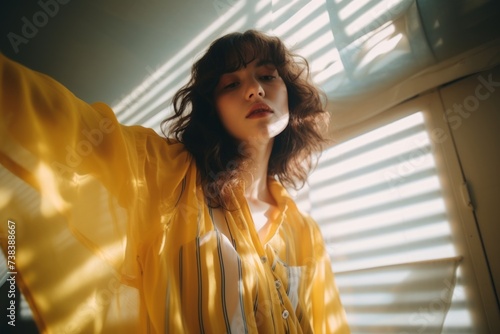 Young beautiful woman with long wavy hair in yellow raincoat standing near window with blinds.