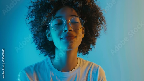 Attractive female student with curly hair wears transparent glasses white t shirt stands against blue background has calm face expression tender smile listens interlocutor with pleasur : Generative AI photo