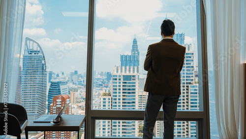 Back view ambitious businessman standing in ornamented office gazing out window to cityscape skyline. Determination and business ambition drive business career toward to bright future