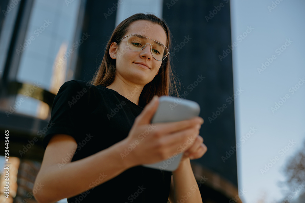 Pretty business woman in eyeglases use mobile phone while standing on modern building background