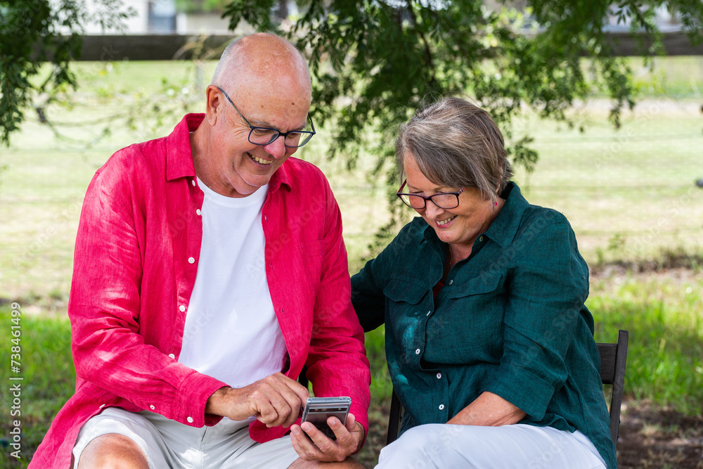 Happy senior couple sitting outside together under tree looking at mobile phone Happy senior couple sitting outside together under tree looking at mobile phone
