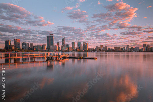 Jetty at South Perth foreshore, Western Australia overlooking city skyline. Water reflections on swan river with cloudy sky, pink, purple, blue hues mirrored on still water, buildings, city, dawn