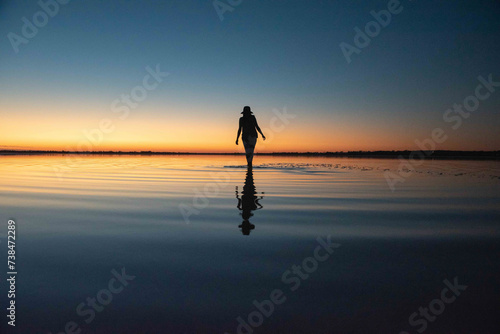 Woman silhouetted against sunset sky walkingin still body of water at Lake Ninan, Western Australia on a summers night. Ripples in water. Concept of peace, stillness, beauty of nature in shallow lake 