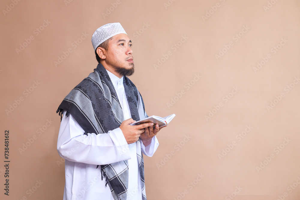 Side view of Asian Muslim man holding Al-Quran while looking away on beige background Side view of Asian Muslim man holding Al-Quran while looking away on beige background