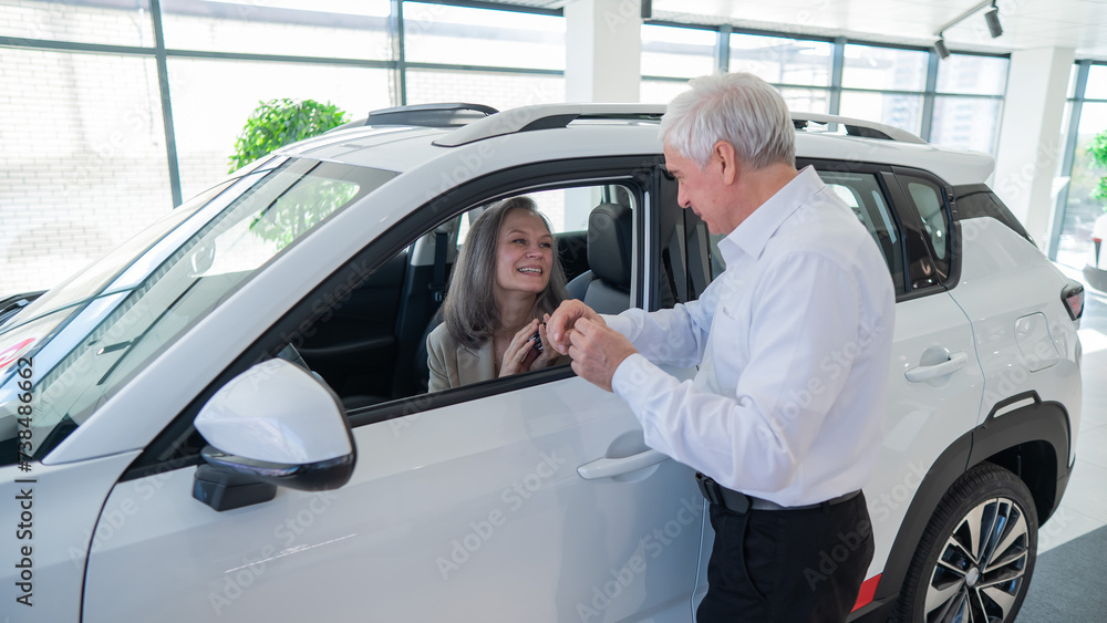 Mature Caucasian woman sits in a new car, an elderly man gives her the keys. Mature Caucasian woman sits in a new car, an elderly man gives her the keys.