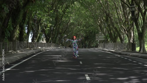 Wallpaper Mural Asian woman in a colored stylish dress with turban walks and dance along the asphalt road under shady trees in the park. Portrait of a woman in motion between trees. Torontodigital.ca