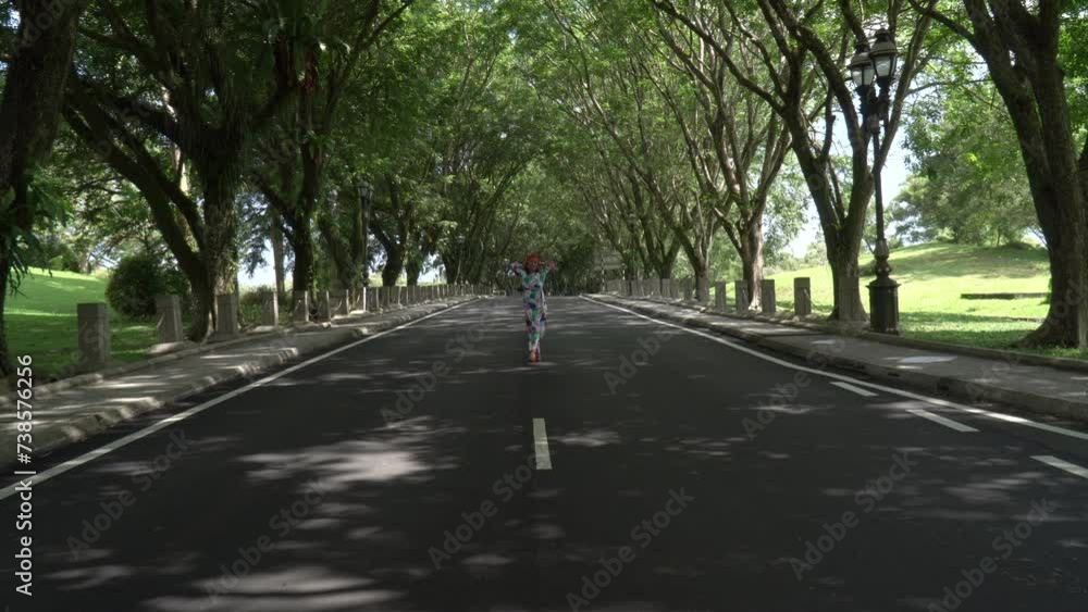 Asian woman in a colored stylish dress with turban walks and dance along the asphalt road under shady trees in the park. Portrait of a woman in motion between trees. Asian woman in a colored stylish dress with turban walks and dance along the asphalt road under shady trees in the park. Portrait of a woman in motion between trees.