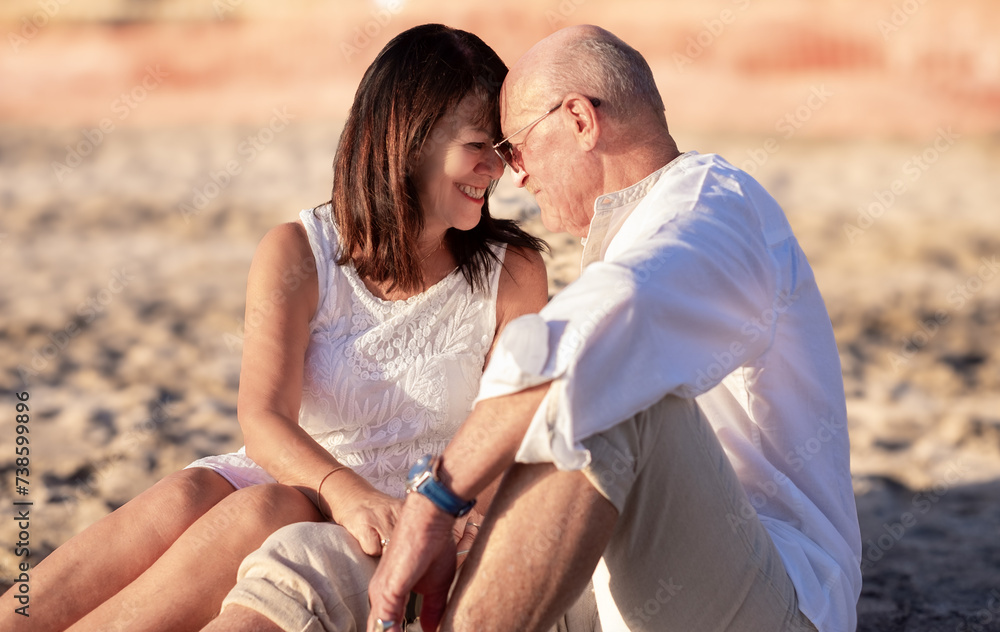 Fototapeta premium Portrait of romantic senior couple sitting on the beach at sunset enjoying vacation and retirement, two elderly people exchange cuddles expressing love and tenderness