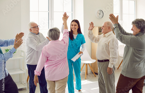 Group of happy lively joyful senior people having fun at retirement home. Good friendly smiling young nurse in uniform with clipboard gives high five to old woman while other patients applaud