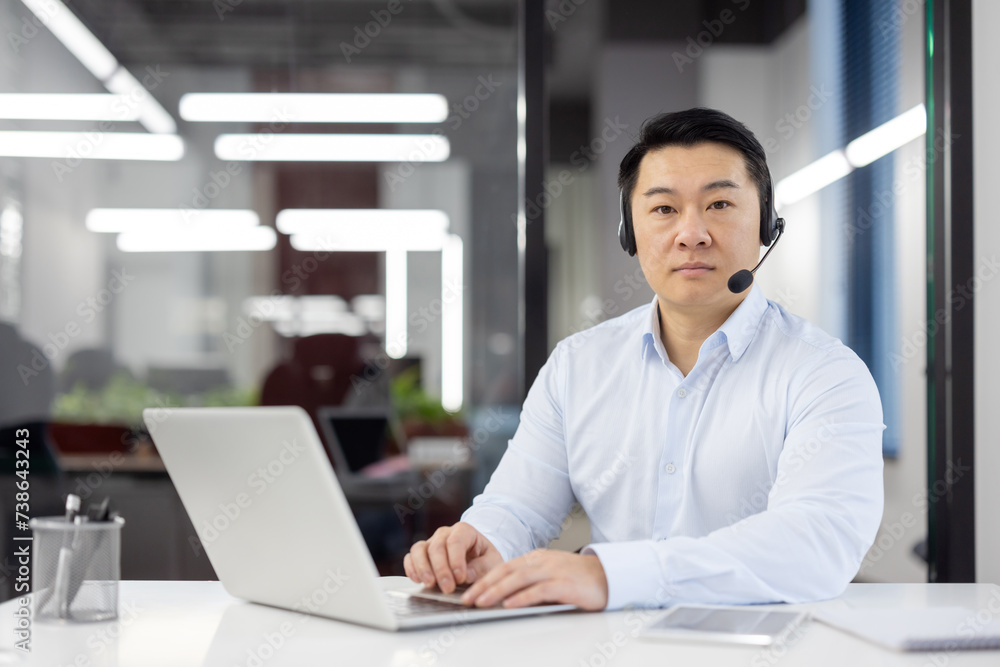 Focused Asian man in smart casual wear using laptop at desk with headset in a contemporary office setting. Focused Asian man in smart casual wear using laptop at desk with headset in a contemporary office setting.