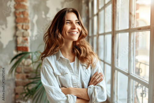 Happy dreamy successful beautiful entrepreneur woman in casual cloth posing in office apartment with hands crossed, looking away, smiling, laughing. Cheerful thoughtful female project owner portrait