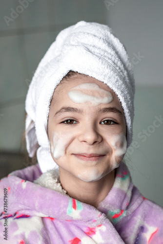 A girl with a bath towel on her head with cleansing foam applied to her face, smiles and looks at the camera.