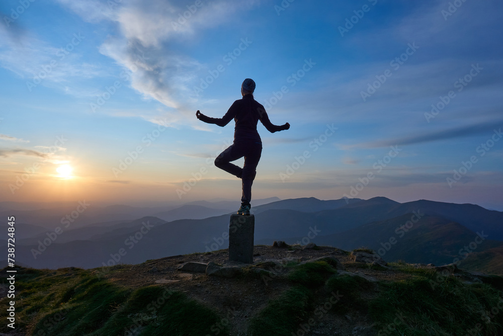 Yoga class in the mountains during sunset. A young girl in dark sportswear stands on a stone in the sun. Beautiful mountain landscape.