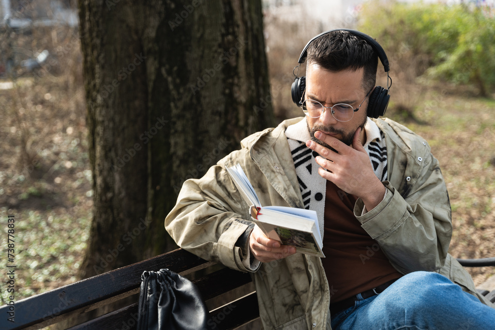 Fototapeta premium Young handsome hipster freelance business man wearing jacket reading a book while sitting outdoors. Businessperson on lunch break listening music on wireless headset and read novel to clear his mind