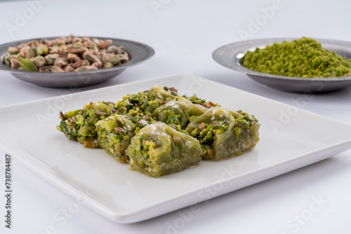 Close up view of traditional Gaziantep cimcik baklava with pistachio on a white plate and ground, raw pistachios in copper plates isolated on white background.