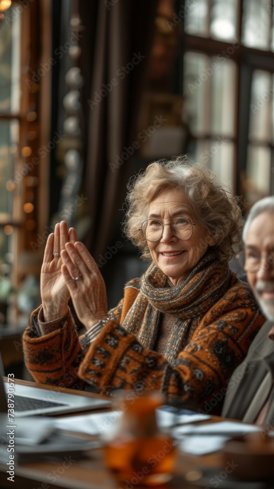 An elderly woman wearing glasses and a scarf smiles and raises her hand in a room