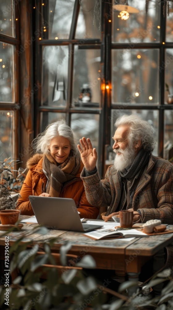 Two elderly people are sitting at a table, smiling, with a laptop, in a cozy cafe