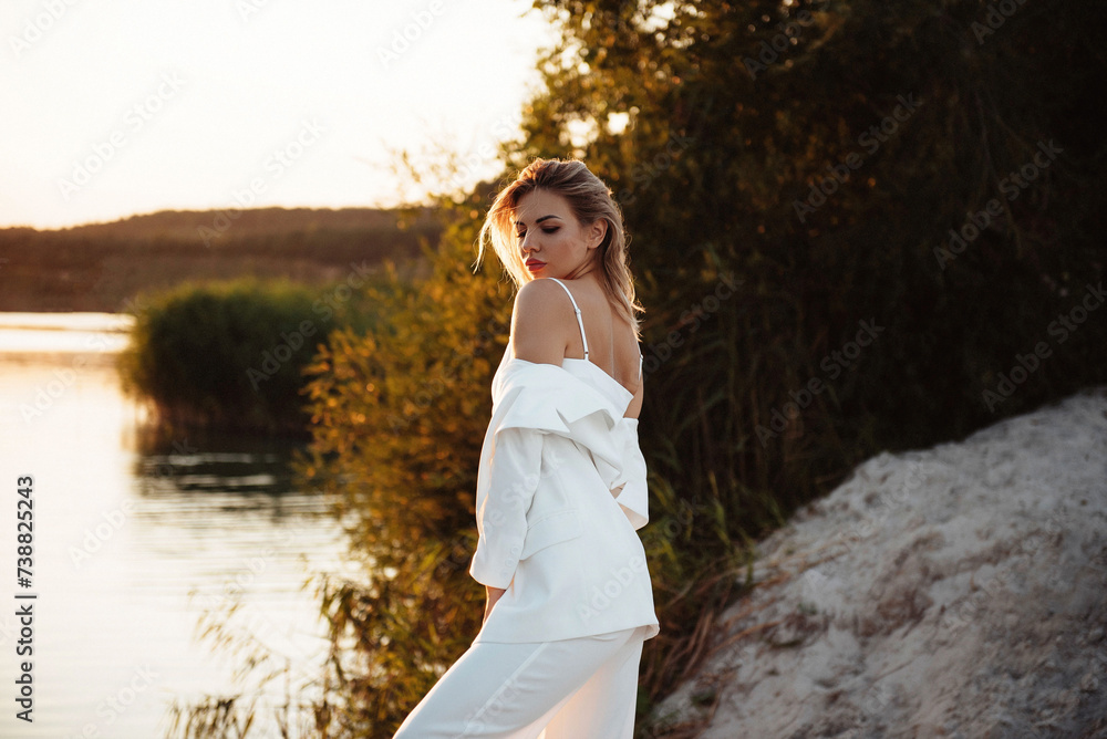Side view of gorgeous blonde in white suit standing near lake during summer sunset. Young woman posing on camera among beautiful nature. Side view of gorgeous blonde in white suit standing near lake during summer sunset. Young woman posing on camera among beautiful nature.