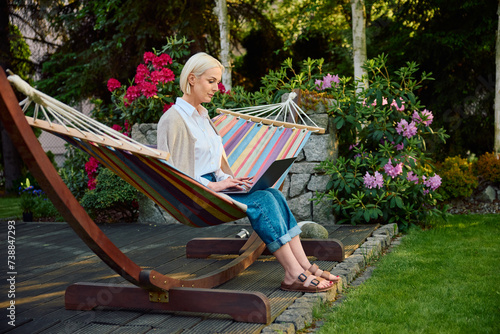Mature woman using laptop sitting on hammock in home backyard patio
